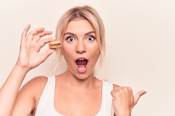 Young blonde woman eating french dessert holding chocolate macaron over white background pointing thumb up to the side smiling happy with open mouth