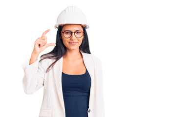 Young beautiful latin girl wearing architect hardhat and glasses smiling and confident gesturing with hand doing small size sign with fingers looking and the camera. measure concept.