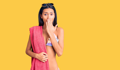 Young beautiful latin girl wearing bikini and sunglasses covering mouth with hand, shocked and afraid for mistake. surprised expression