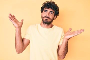 Handsome young man with curly hair and bear wearing casual tshirt over yellow background clueless...
