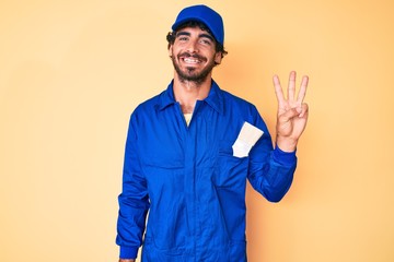 Handsome young man with curly hair and bear wearing builder jumpsuit uniform showing and pointing up with fingers number three while smiling confident and happy.