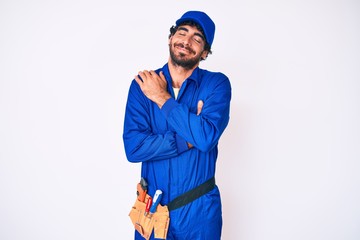 Handsome young man with curly hair and bear weaing handyman uniform hugging oneself happy and...