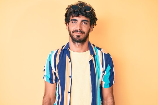 Handsome young man with curly hair and bear wearing summer shirt and sunglasses with serious expression on face. simple and natural looking at the camera.