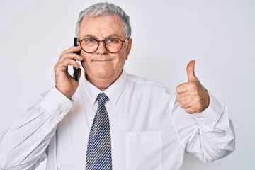 Senior grey-haired man wearing business clothes having conversation talking on the smartphone smiling happy and positive, thumb up doing excellent and approval sign