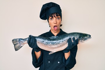 Beautiful brunettte woman professional chef holding fresh salmon fish in shock face, looking...