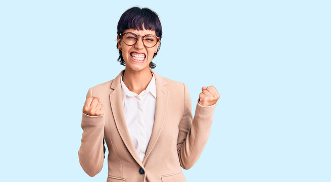 Young brunette woman with short hair wearing business jacket and glasses excited for success with arms raised and eyes closed celebrating victory smiling. winner concept.