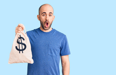 Young handsome man holding money bag with dollar symbol scared and amazed with open mouth for surprise, disbelief face
