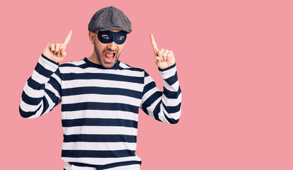 Young handsome man wearing burglar mask smiling amazed and surprised and pointing up with fingers and raised arms.