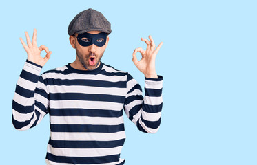 Young handsome man wearing burglar mask looking surprised and shocked doing ok approval symbol with fingers. crazy expression