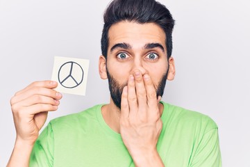 Young handsome man with beard holding peace symbol reminder covering mouth with hand, shocked and...