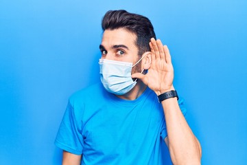 Young handsome man with beard wearing medical mask smiling with hand over ear listening and hearing to rumor or gossip. deafness concept.