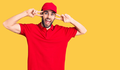Young handsome man with beard wearing delivery uniform doing peace symbol with fingers over face, smiling cheerful showing victory