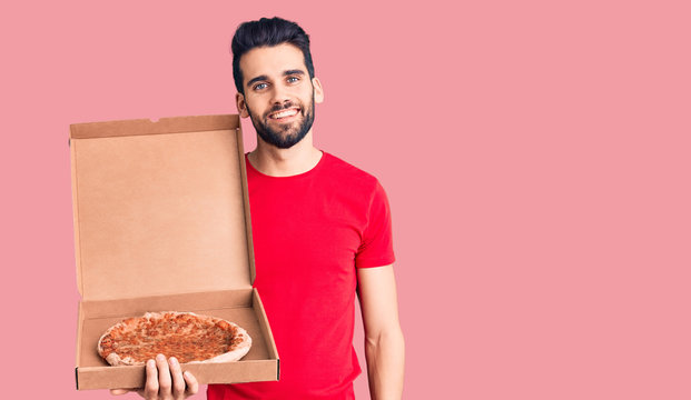 Young handsome man with beard holding delivery cardboard with italian pizza looking positive and happy standing and smiling with a confident smile showing teeth