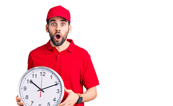 Young handsome man with beard wearing delivery uniform holding big clock scared and amazed with open mouth for surprise, disbelief face