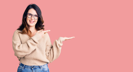 Beautiful young brunette woman wearing casual clothes and glasses amazed and smiling to the camera while presenting with hand and pointing with finger.