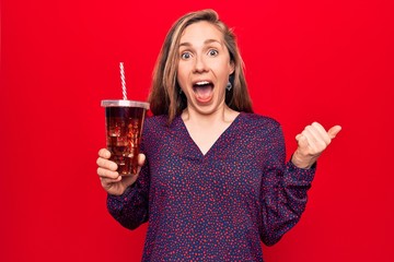 Young beautiful blonde woman drinking a fresh glass of soda pointing thumb up to the side smiling happy with open mouth