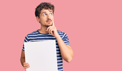 Young handsome man with curly hair holding blank empty banner serious face thinking about question...