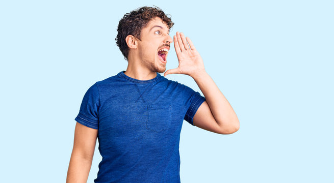 Young handsome man with curly hair wearing casual clothes shouting and screaming loud to side with hand on mouth. communication concept.