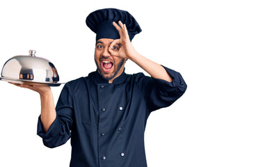 Young hispanic man wearing cooker uniform holding tray smiling happy doing ok sign with hand on eye looking through fingers