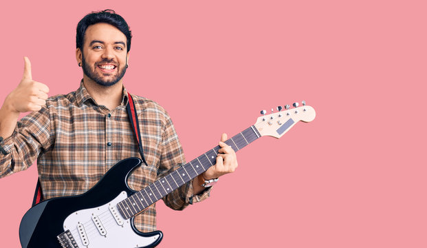 Young hispanic man playing electric guitar smiling happy and positive, thumb up doing excellent and approval sign