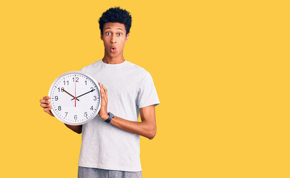 Young african american man holding big clock scared and amazed with open mouth for surprise, disbelief face