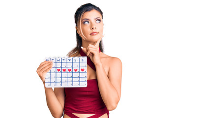 Young beautiful woman holding heart calendar serious face thinking about question with hand on chin, thoughtful about confusing idea