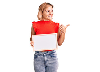 Young blonde woman holding empty white chalkboard pointing thumb up to the side smiling happy with open mouth