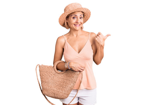 Young blonde woman wearing summer hat holding bag pointing thumb up to the side smiling happy with open mouth