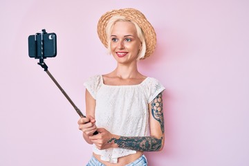Young blonde woman with tattoo taking a selfie photo with smartphone looking positive and happy standing and smiling with a confident smile showing teeth
