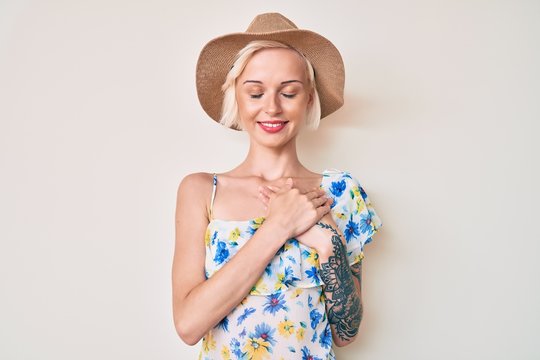 Young blonde woman with tattoo wearing summer hat smiling with hands on chest with closed eyes and grateful gesture on face. health concept.