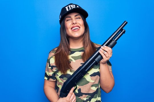 Young beautiful hunter woman wearing camouflage t-shirt and usa cap holding shotgun looking positive and happy standing and smiling with a confident smile showing teeth
