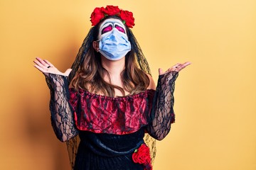 Obraz premium Young woman wearing day of the dead costume wearing medical mask celebrating mad and crazy for success with arms raised and closed eyes screaming excited. winner concept