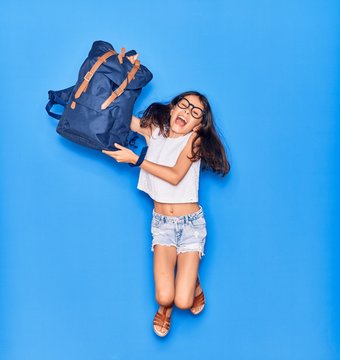 Adorable hispanic student child girl wearing glasses smiling happy. Jumping with smile on face holding backpack over isolated blue background