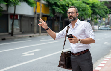 Confidence Caucasian business man office worker carry leather bag and using mobile app on smartphone call a cab in downtown district. Handsome beard guy passenger hail a taxi on city sidewalk.
