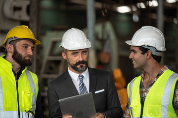 Factory manager in suit using laptop and talking with industrial engineer ,technician man. They wear helmet or hardhat with safety jackets inside industry manufacturing Facility/Discussion  working