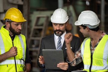 Factory manager in suit using laptop and talking with industrial engineer ,technician man. They wear helmet or hardhat with safety jackets inside industry manufacturing Facility/Discussion  working