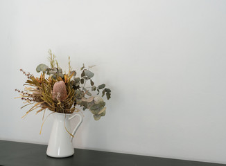 Close up of dried Australian native flowers in white jug on dark bench against wall (selective focus)