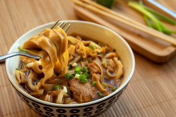 In the foreground a wide yellow noodle on a fork. In the background, a plate of Asian noodle soup with dark broth, chunks of beef and greens in a deep round plate. The plate is on a bamboo tablecloth. © Vadim