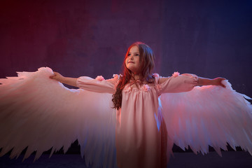 Naklejka premium Small young model girl with white wings posing and dancing in dark black studo during photoshoot with flour or dust and colour light. Actress in performance about struggle between good and evil.