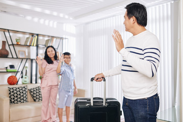 Mature Asian man with suitcase waving to his wife and preteen son when leaving for business trip