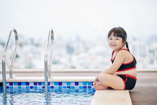 Cheerful little Vietnamese girl sitting on edge of swimming pool and looking at camera