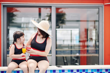 Hugging mother and daughter sitting on pool edge and drinking fresh fruit cocktails