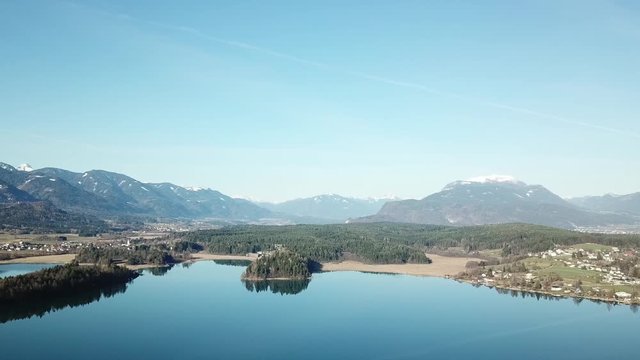 A drone flight above the Faaker See in Austria. There are high Alpine ranges around, reflecting in the crystal clear and calm surface of the lake. Small villages around the lake. Snow capped mountains