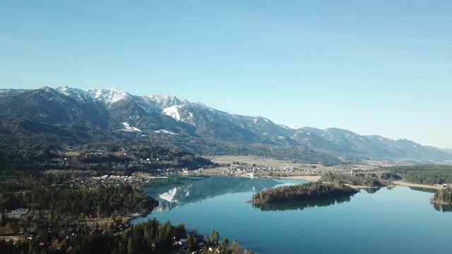 A drone flight above the Faaker See in Austria. There are high Alpine ranges around, reflecting in the crystal clear and calm surface of the lake. Small villages around the lake. Snow capped mountains