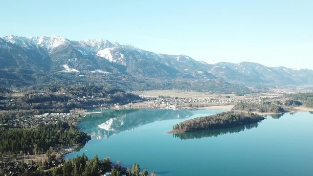 A drone flight above the Faaker See in Austria. There are high Alpine ranges around, reflecting in the crystal clear and calm surface of the lake. Small villages around the lake. Snow capped mountains