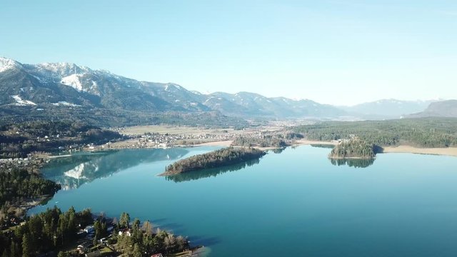 A drone flight above the Faaker See in Austria. There are high Alpine ranges around, reflecting in the crystal clear and calm surface of the lake. Small villages around the lake. Snow capped mountains