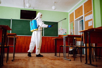 Man sprays disinfection liquid over the desks at the classroom before the school season. Sanitary worker wearing protective suit cleans the auditorium. Students and pupils health care concept.