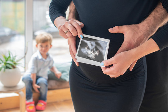 A pregnant woman in a black dress holds an ultrasound image of her baby and her husband supports her stomach, with a small child sitting in the background: the concept of women's health