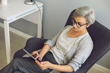 Online work education communication. Senior woman with glasses uses tablet at home.