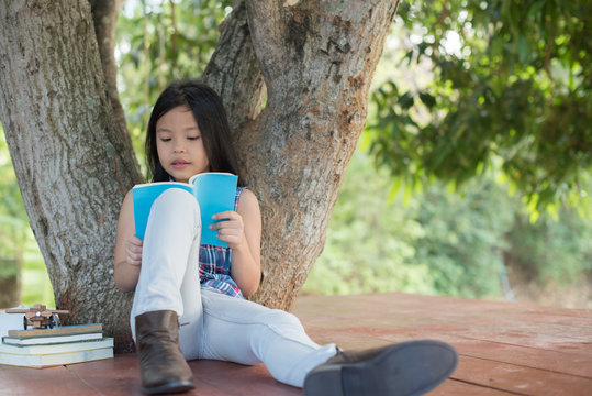 little asian girl reading a book under big tree with hat, book, and model plane beside. children and science. blurred background. learning the imagination and dreams of rural child. studying at home.